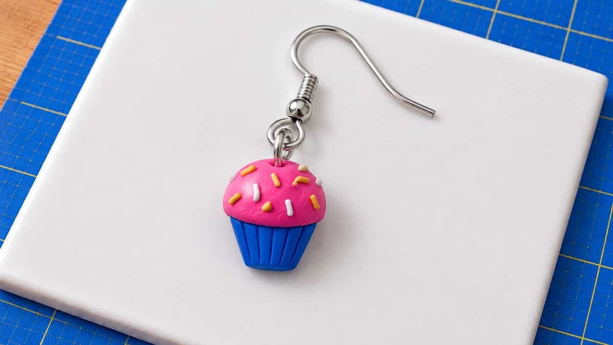 A miniature blue polymer clay cupcake wrapper supports a textured pink dome with yellow and white clay sprinkles, connected to a silver eye-pin loop, jump ring and ear wire.