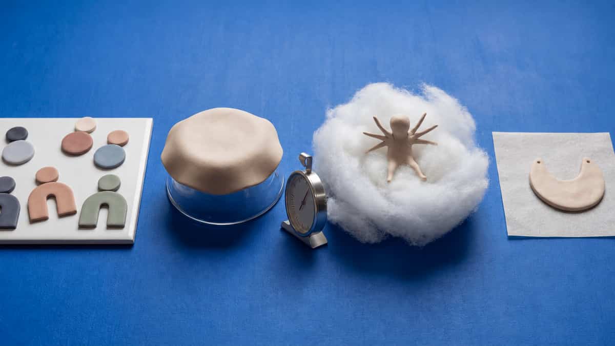 Baking supports on a blue background: flat clay shapes on a glazed tile, a clay dome over an upturned glass bowl, a dial oven thermometer, a clay figure in a fiberfill nest and a crescent pendant on flat parchment.