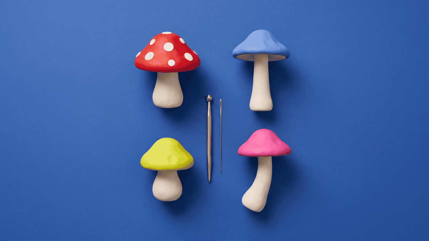 Red toadstool polymer clay mushroom with white spots beside blue, yellow and pink clay mushrooms on cream stems, with a ball tool and a needle tool on a blue background