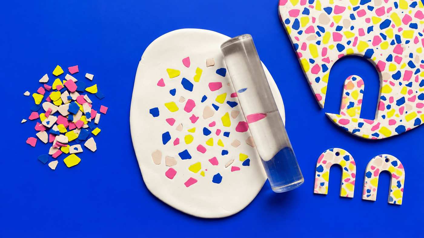 Pink, yellow, blue and pale pink clay chips beside a white slab and clear acrylic roller, with an arch cutout in a terrazzo slab and two terrazzo arch pieces on a blue surface.