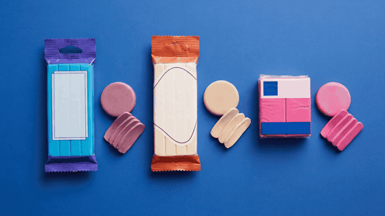 Three unbranded packs of polymer clay on a cobalt blue background: a turquoise bar in a purple wrapper, a cream bar in an orange wrapper and a square pink block, each with a rolled disc and a few cut slices of its clay beside it