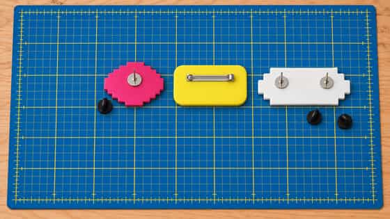 Three polymer clay brooches face down on a blue cutting mat: a pink one with a tie-tack pin and its black clutch, a yellow one with a bar pin across the top third, and a wide white one with two tie-tacks at the same height.