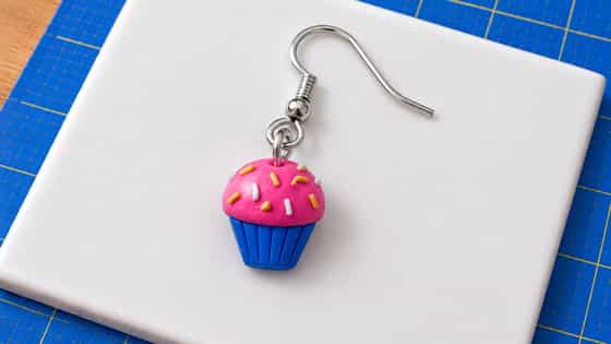 A miniature blue polymer clay cupcake wrapper supports a textured pink dome with yellow and white clay sprinkles, connected to a silver eye-pin loop, jump ring and ear wire.