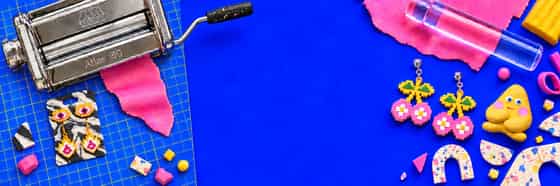Bright pink clay, a roller, clay-conditioning machine and colourful polymer clay jewellery arranged across an electric-blue studio surface.