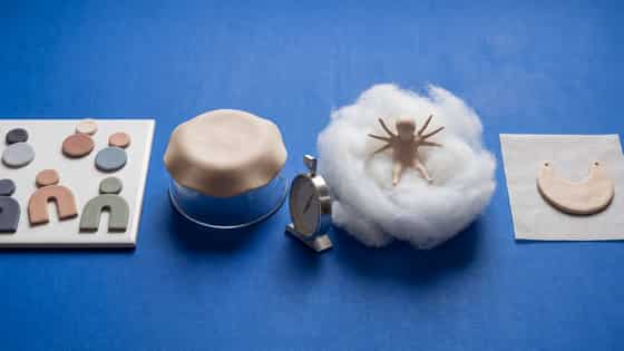 Baking supports on a blue background: flat clay shapes on a glazed tile, a clay dome over an upturned glass bowl, a dial oven thermometer, a clay figure in a fiberfill nest and a crescent pendant on flat parchment.