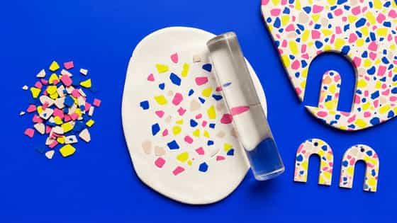 Pink, yellow, blue and pale pink clay chips beside a white slab and clear acrylic roller, with an arch cutout in a terrazzo slab and two terrazzo arch pieces on a blue surface.