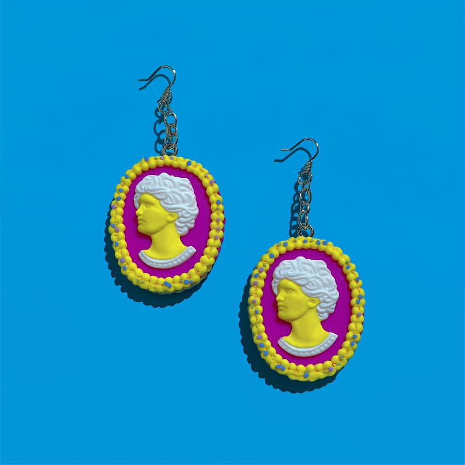 Pair of polymer clay cameo drop earrings on a bright blue background: fuchsia ovals with raised yellow profiles, white hair and collars, and beaded yellow borders flecked with blu…