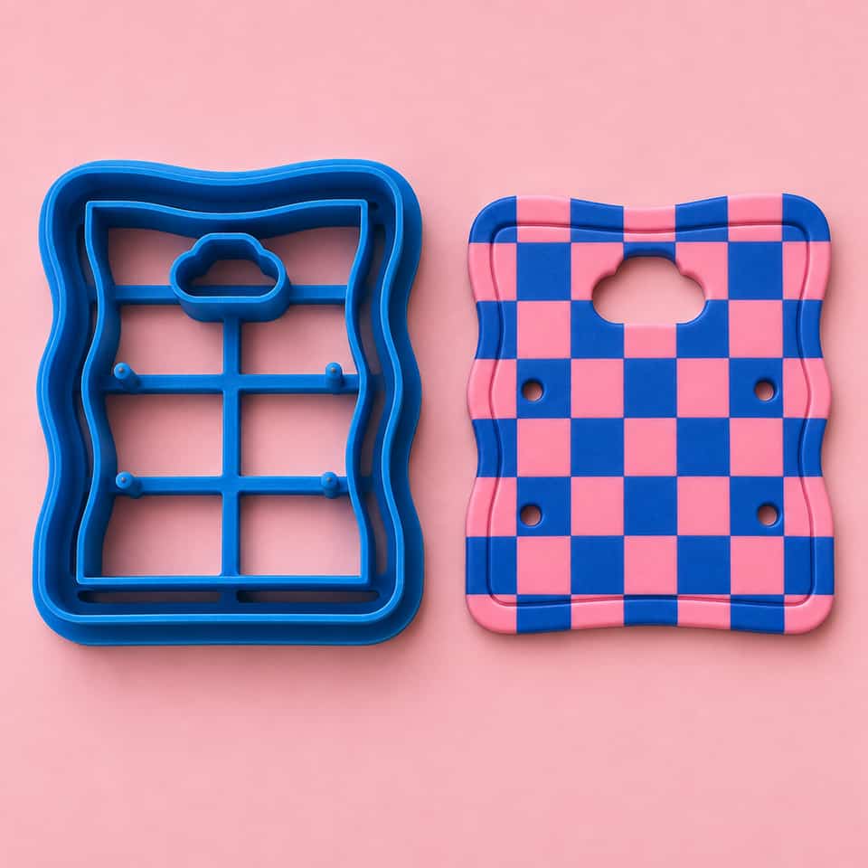 A blue 3D-printed card cutter beside a pink-and-blue checker-pattern clay card with its hanger and four round holes open.