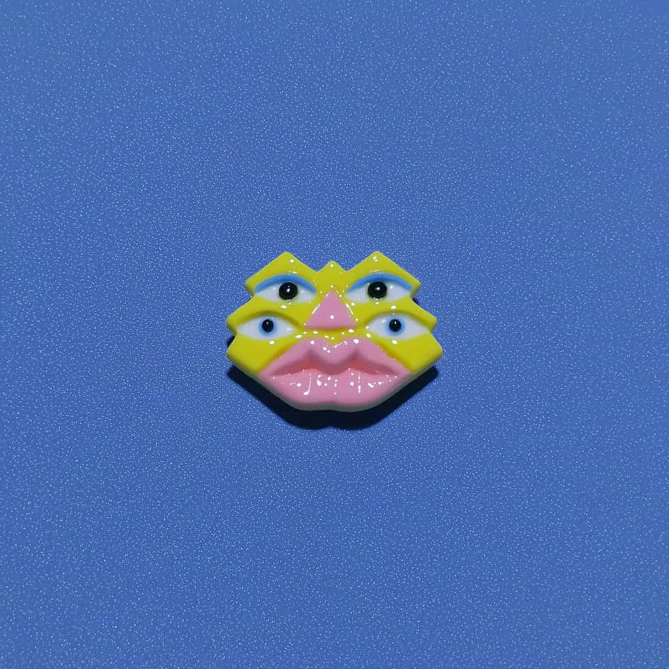 Glossy polymer clay face brooch: a lemon yellow face with zigzag points and notches, four white almond eyes with black pupils, pale blue lids on the top pair and blue rings on the…