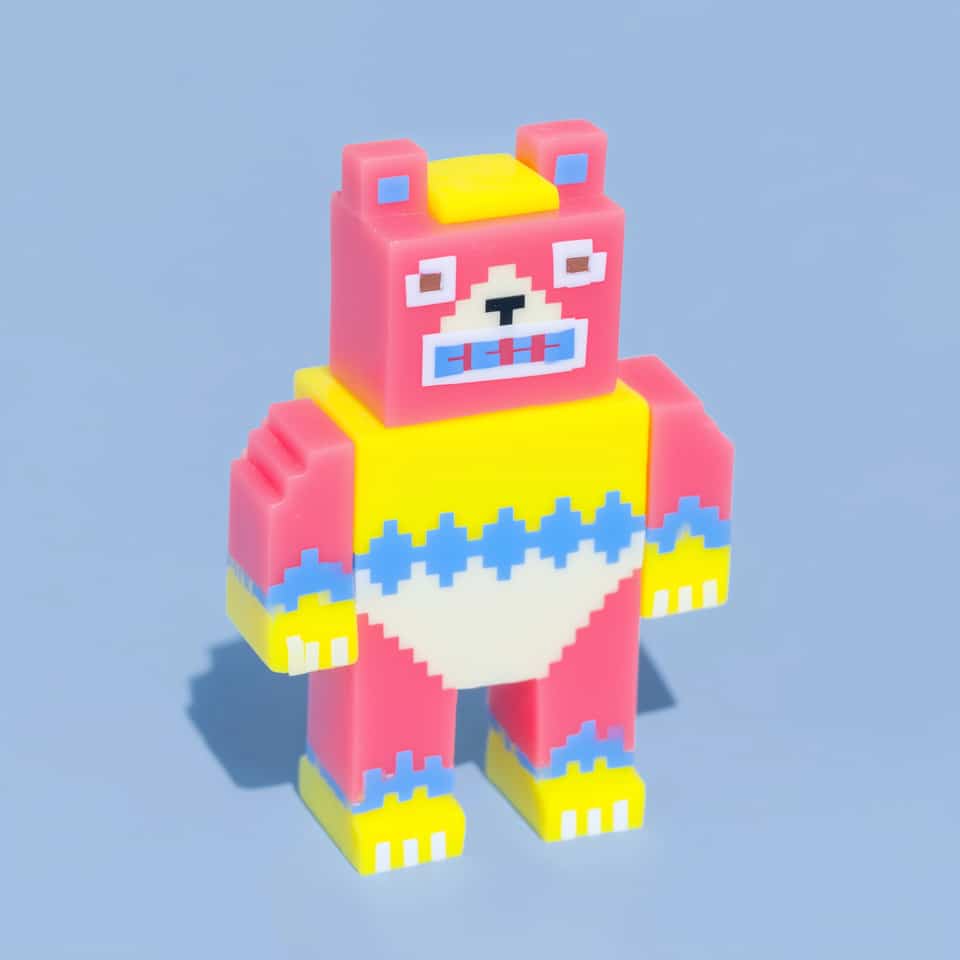 Blocky pink and yellow pixel bear figurine standing upright: pink head with blue-centred ears, square white eyes with brown pupils and a framed blue mouth, yellow chest with a blu…