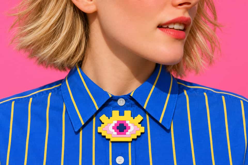 A layered Pixel Eye brooch sits between the collars of a striped shirt.
