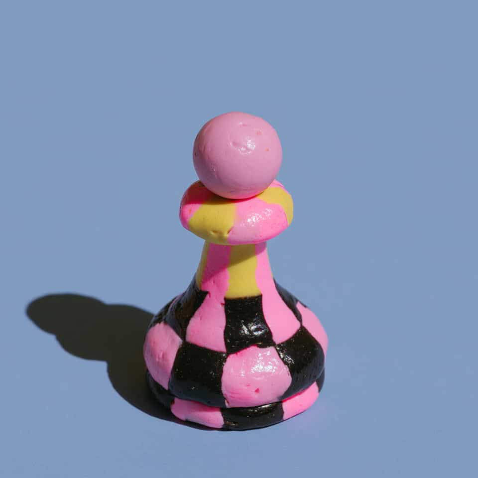Polymer clay chess pawn about 5 cm tall with a plain pink ball head, a pink and yellow collar, a pink and yellow striped stem and a body and base in pink and black checks, with a…