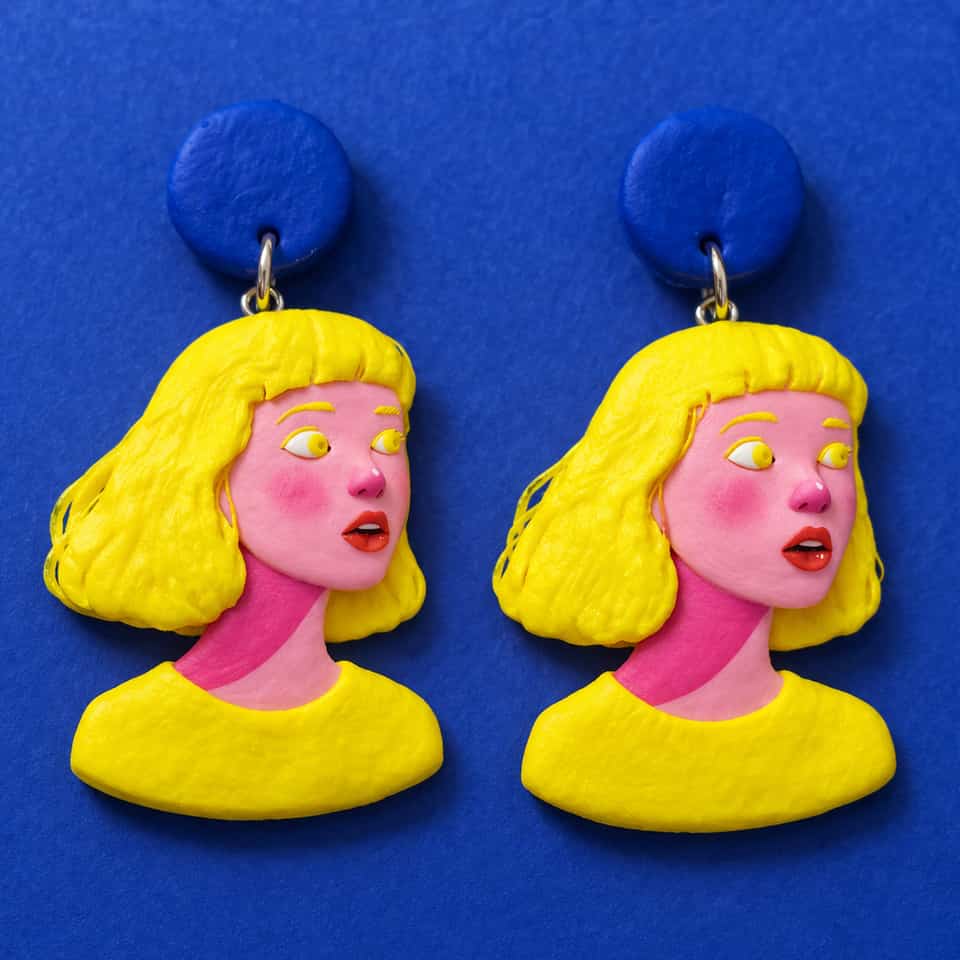 Pair of polymer clay portrait drop earrings: two identical girls with textured yellow bobs, pink faces with blushed cheeks, yellow eyes glancing sideways, open red lips and yellow…