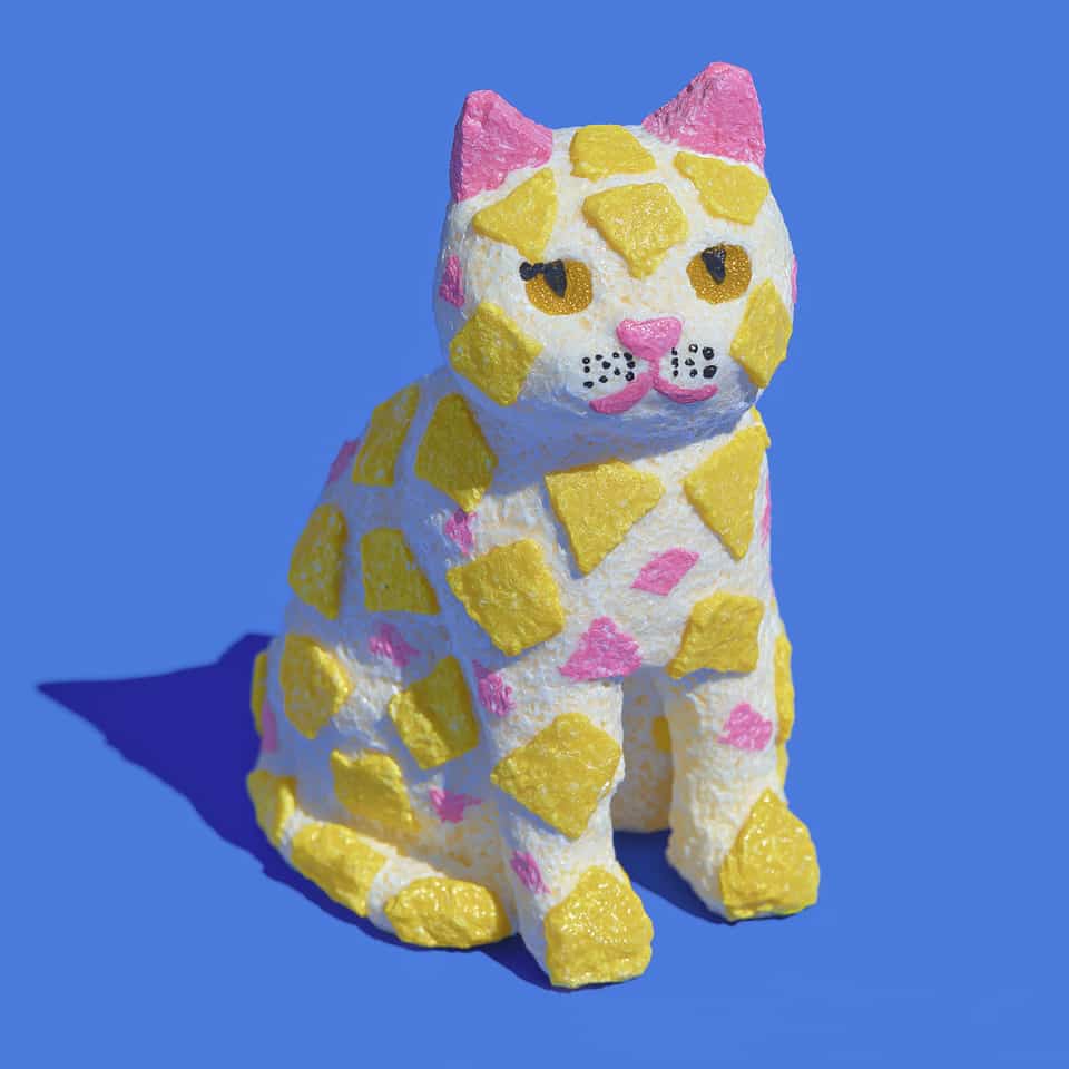 Seated cream-white polymer clay cat figurine with a rough stone texture, covered in angular yellow and small pink terrazzo chips, with solid pink ears, amber almond eyes with blac…