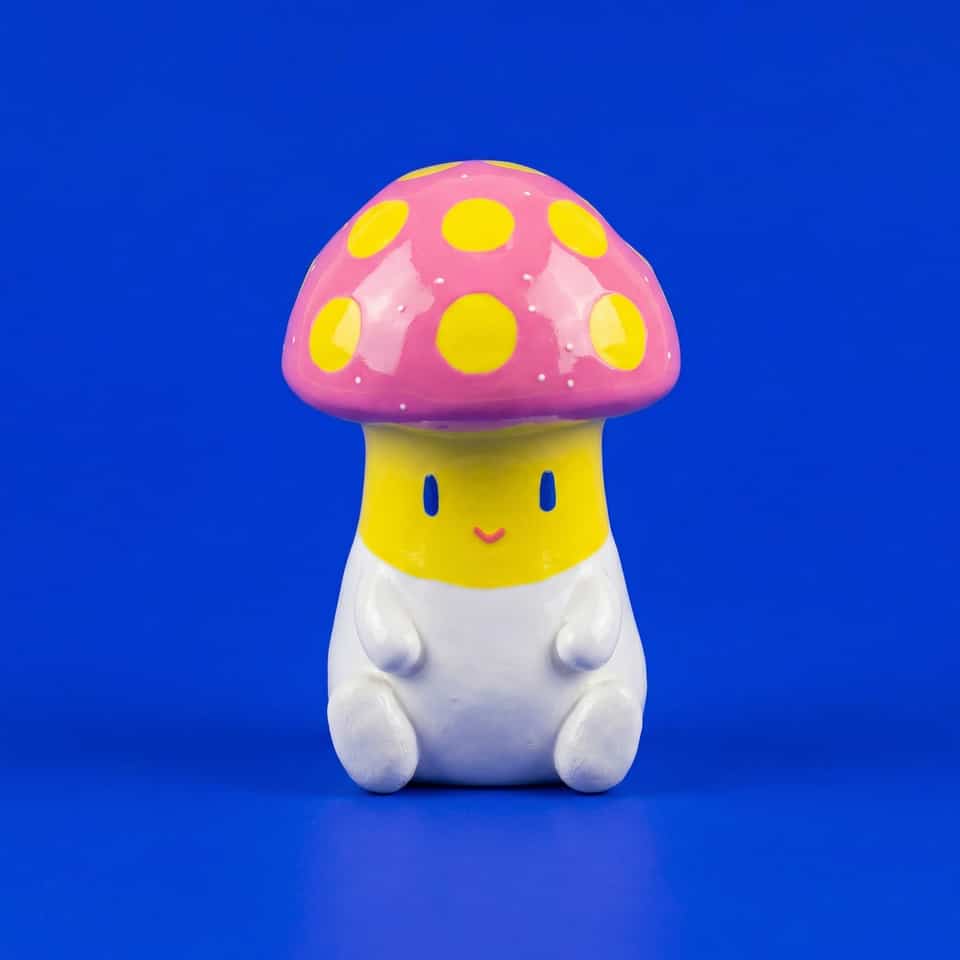 Polymer clay mushroom figurine with a glossy pink cap covered in flat yellow spots and tiny white specks, a yellow face band with tall blue eyes and a small coral smile, and a whi…