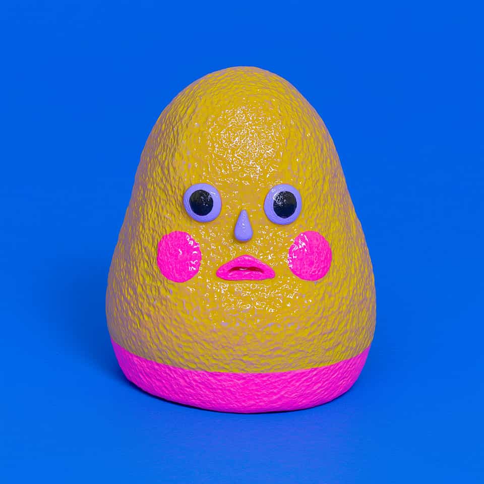 Tall egg-shaped yellow polymer clay blob figurine with a coarse pebbled texture and a hot-pink band round the base, lilac ring eyes with black pupils, a small lilac teardrop nose,…