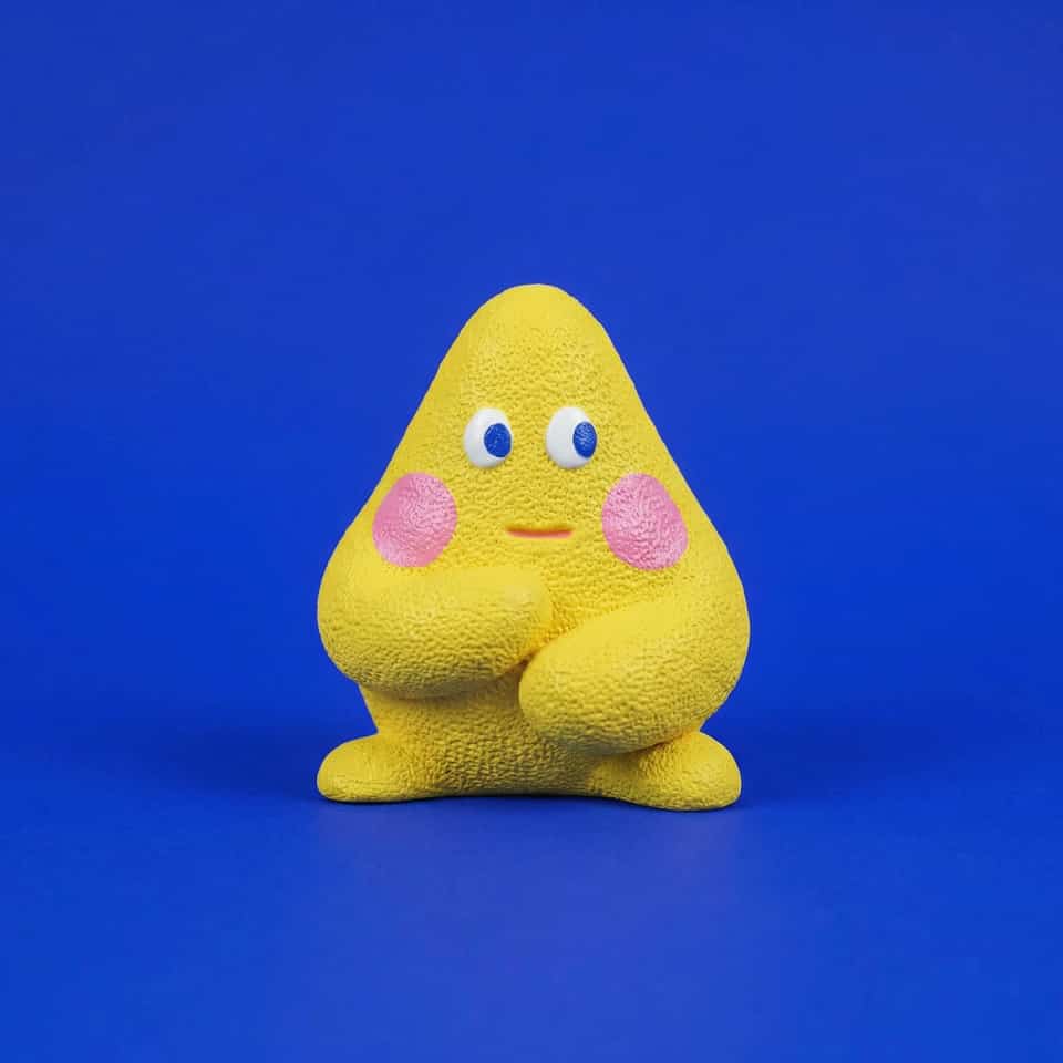 Yellow polymer clay blob figurine with a pebbled texture, chunky arms folded across its belly, pink cheeks, white eyes with blue pupils glancing sideways and a small coral mouth.