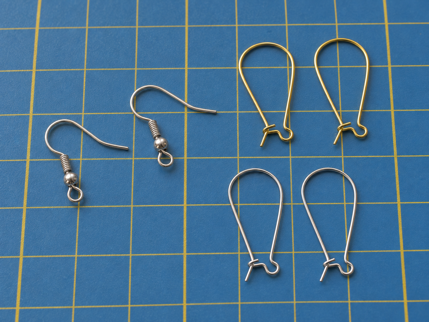 Silver fish-hook ear wires with ball and coil detail beside gold and silver kidney ear wires on a blue cutting mat.