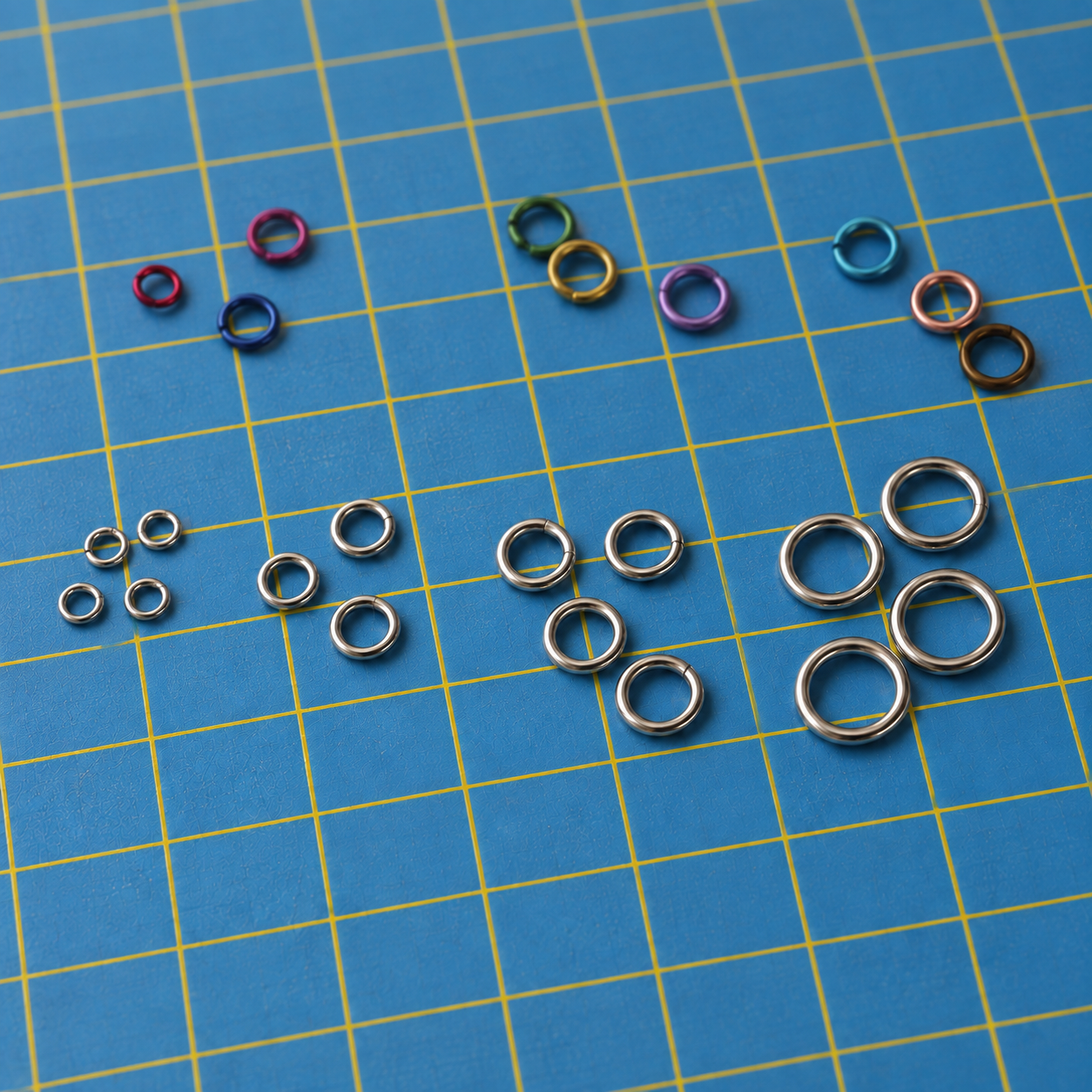 Silver-tone open jump rings in four sizes arranged in rows on a blue cutting mat, with a few small coloured rings above them.