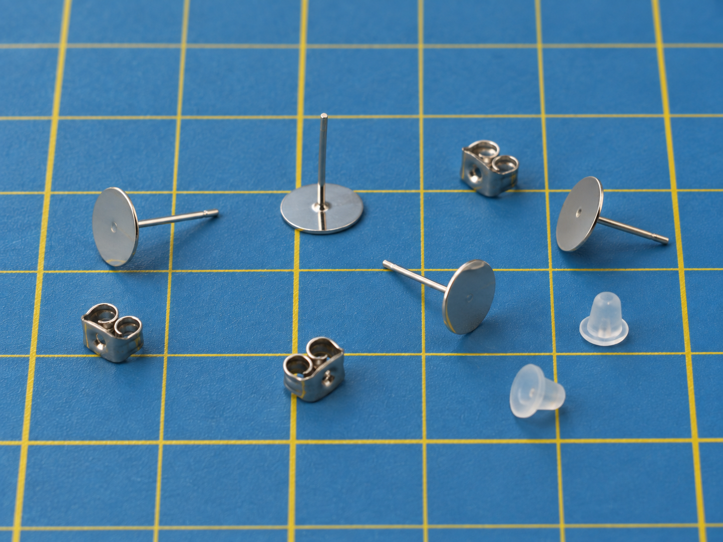 Silver-tone flat-pad earring posts with metal butterfly backs and two clear silicone backs on a blue cutting mat.