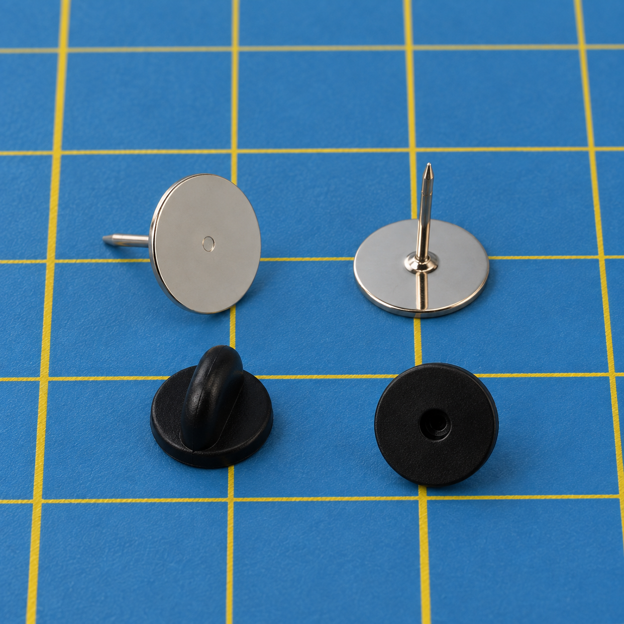 Two silver flat-pad tie-tack pins on a blue cutting mat with a yellow grid, one showing the flat face of its round pad and one standing pad-down with its pointed post up, with two black clutches below them, one seen from the side and one showing the hole the post goes into