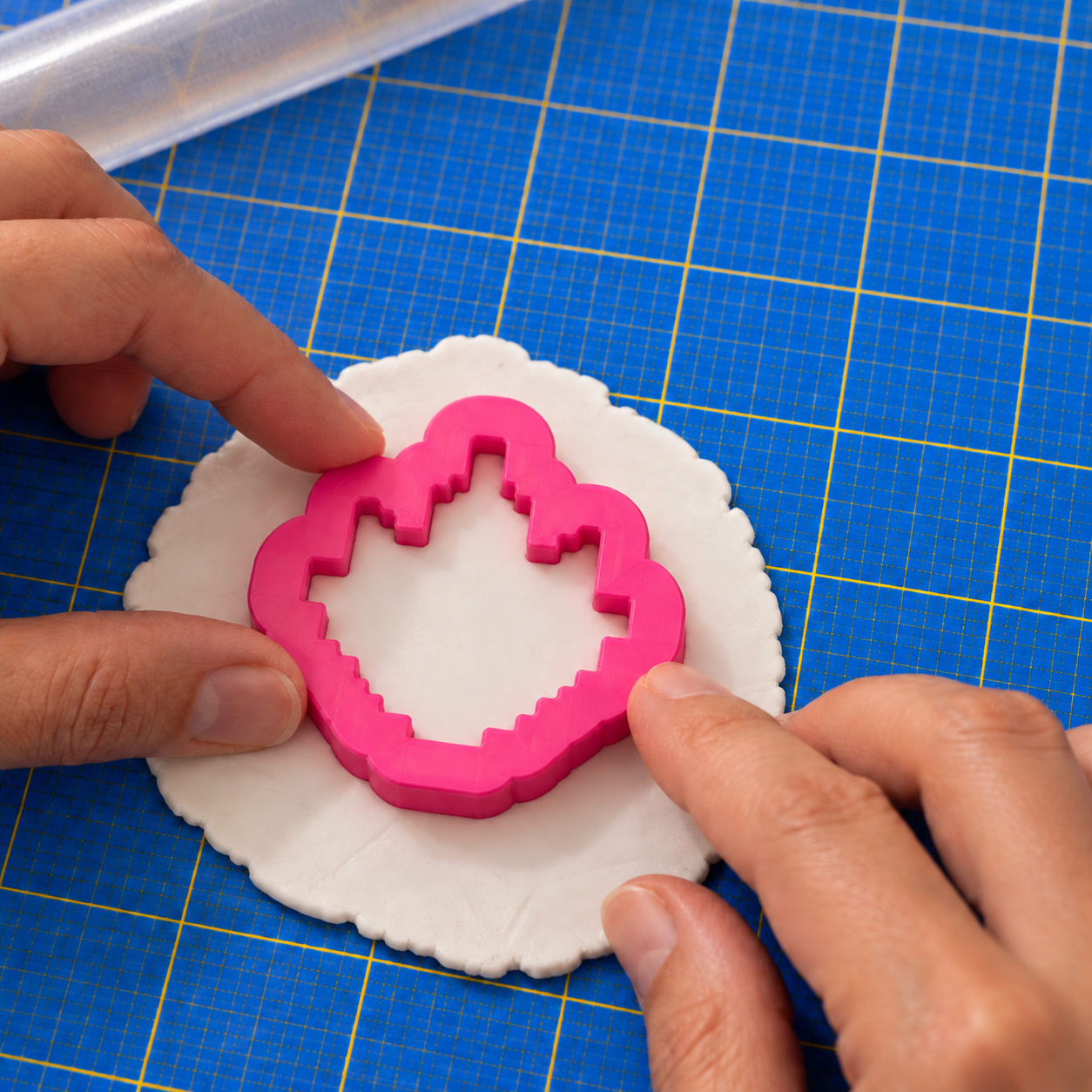 Two hands pressing a pink printed pixel-flame cutter into a round sheet of white polymer clay on a blue grid cutting mat.
