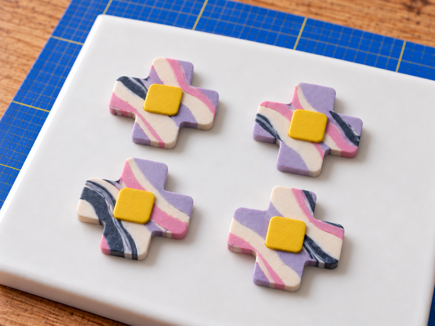 Four small cross-shaped clay flower fronts with yellow square centres; lilac, pink, cream and charcoal ribbons form a different pattern in each flower.