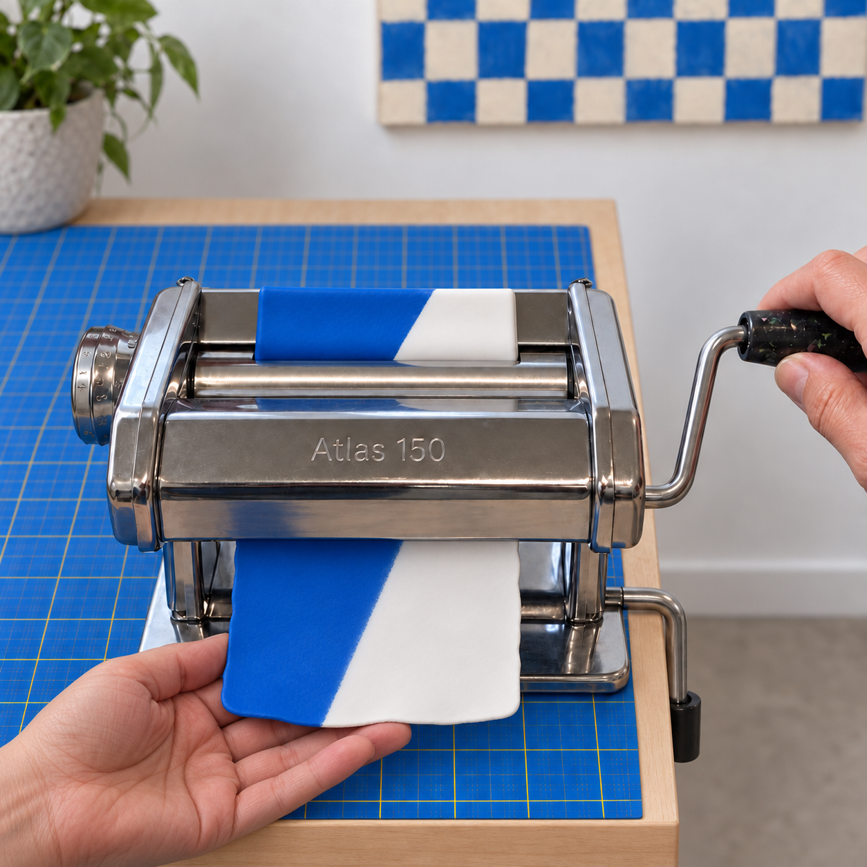 A hand feeding the joined blue and white clay rectangle into the rollers of a chrome Atlas 150 pasta machine on a blue mat.