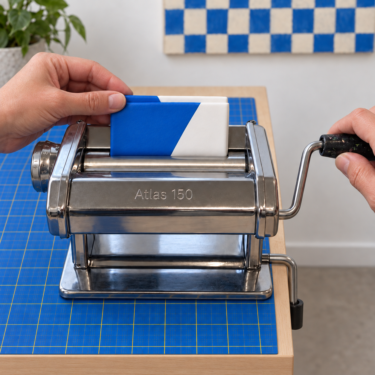 A hand guiding a folded blue and white clay sheet, folded edge downward, into the rollers of a pasta machine while the other hand turns the crank.