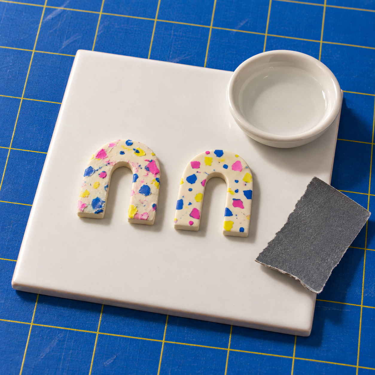 Two baked terrazzo arches on a white tile beside a small dish of water and a torn strip of wet-and-dry sandpaper: the left arch has a hazy, smeared surface; the right arch is sanded flat with crisp chip edges.