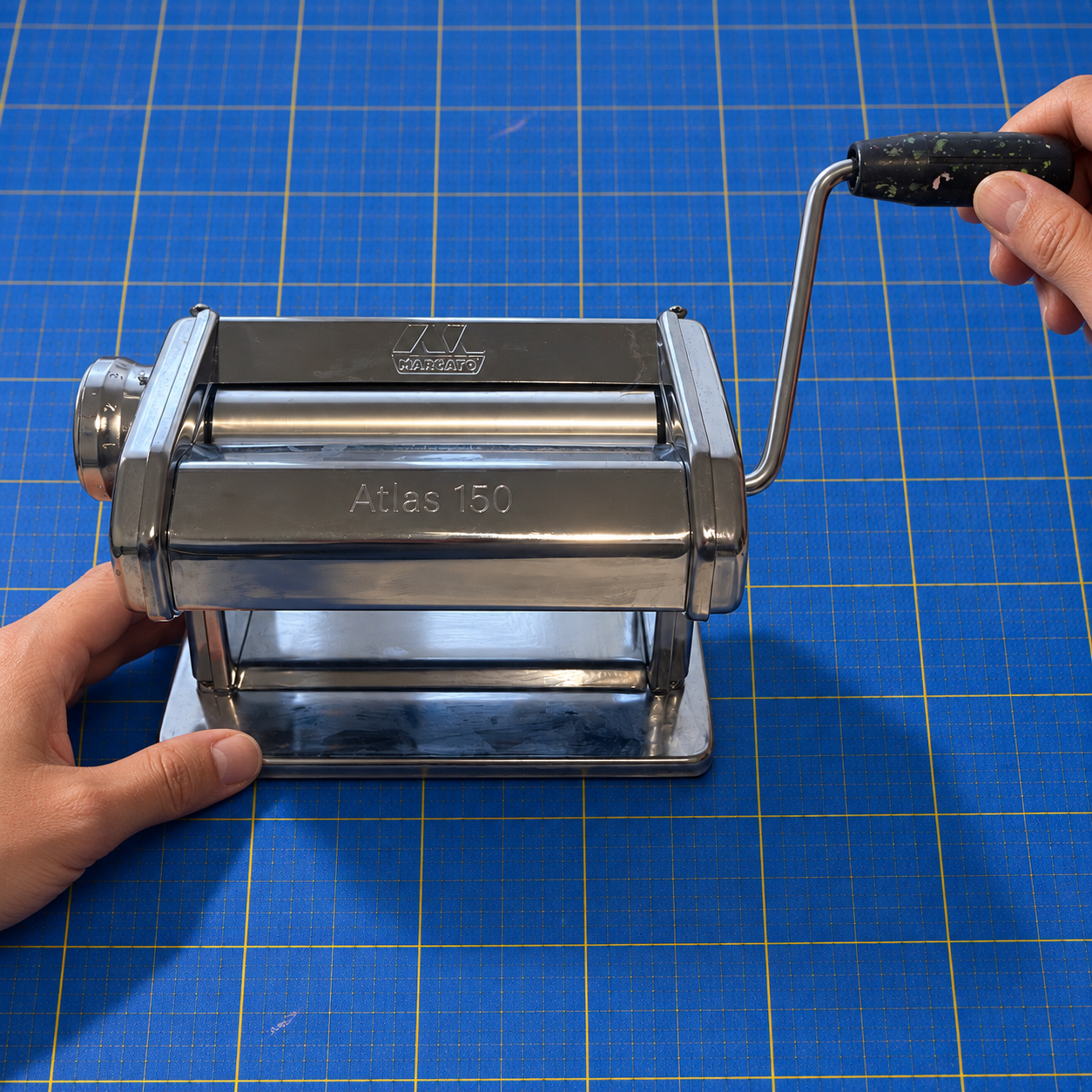 Two hands working a chrome Atlas 150 pasta machine on a blue cutting mat, one steadying the body and one turning the handle.