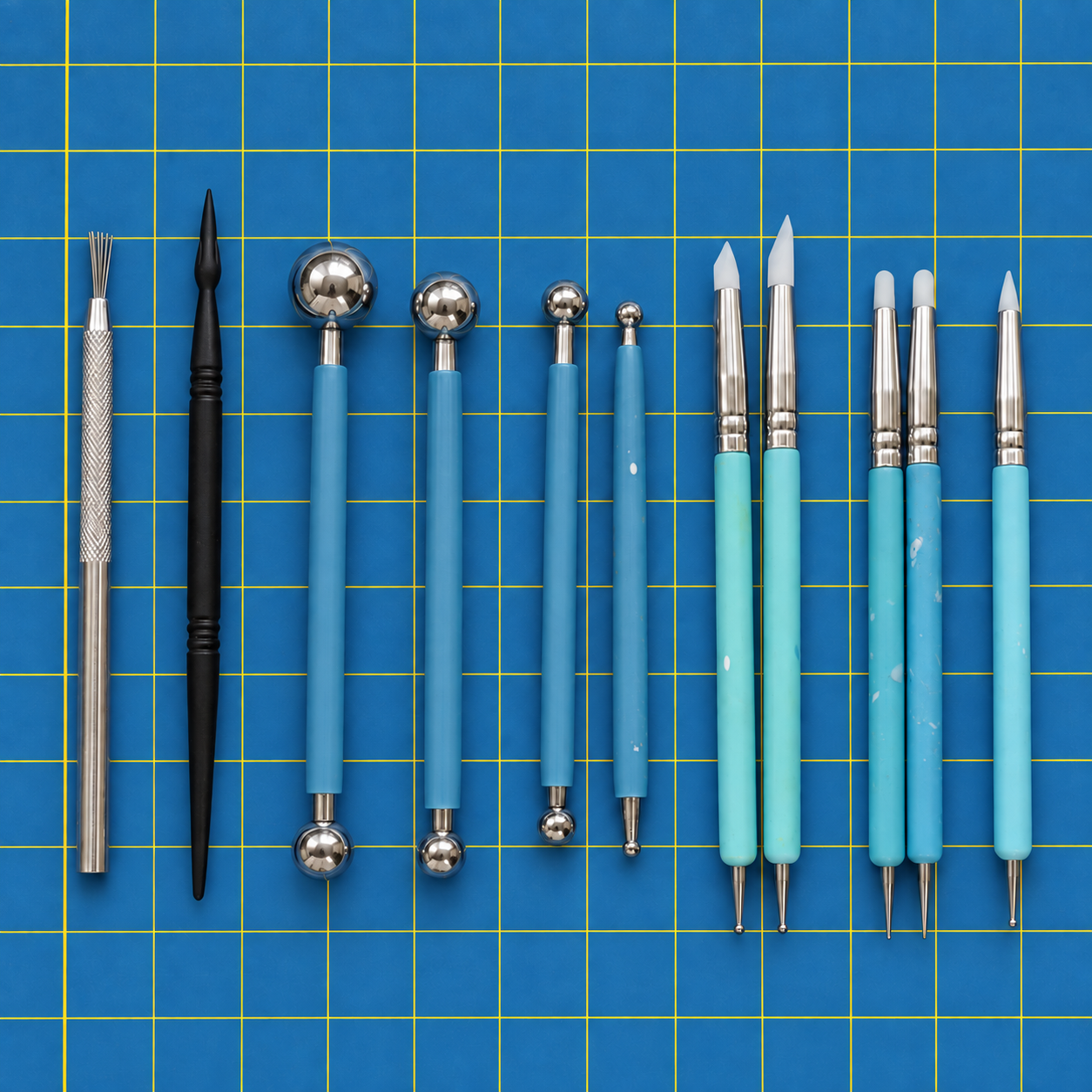 A row of double-ended sculpting tools with blue handles on a blue cutting mat: ball styluses in several sizes, silicone shaper tips, needle points, a wire texture tool and a black wedge tool.