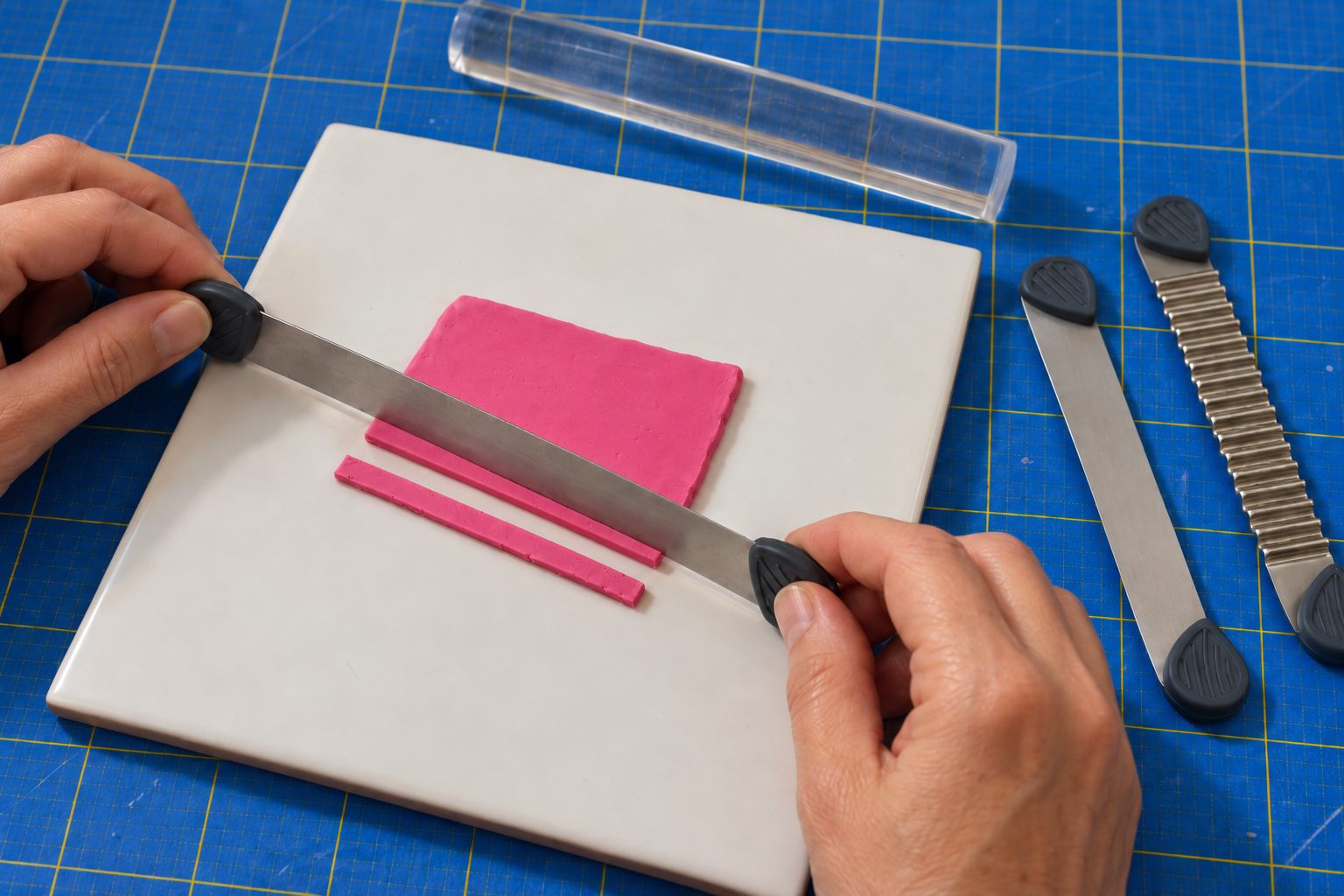 Two hands guiding a straight tissue blade through a pink clay slab on a white tile, with an acrylic roller and a wavy blade nearby on a blue cutting mat.