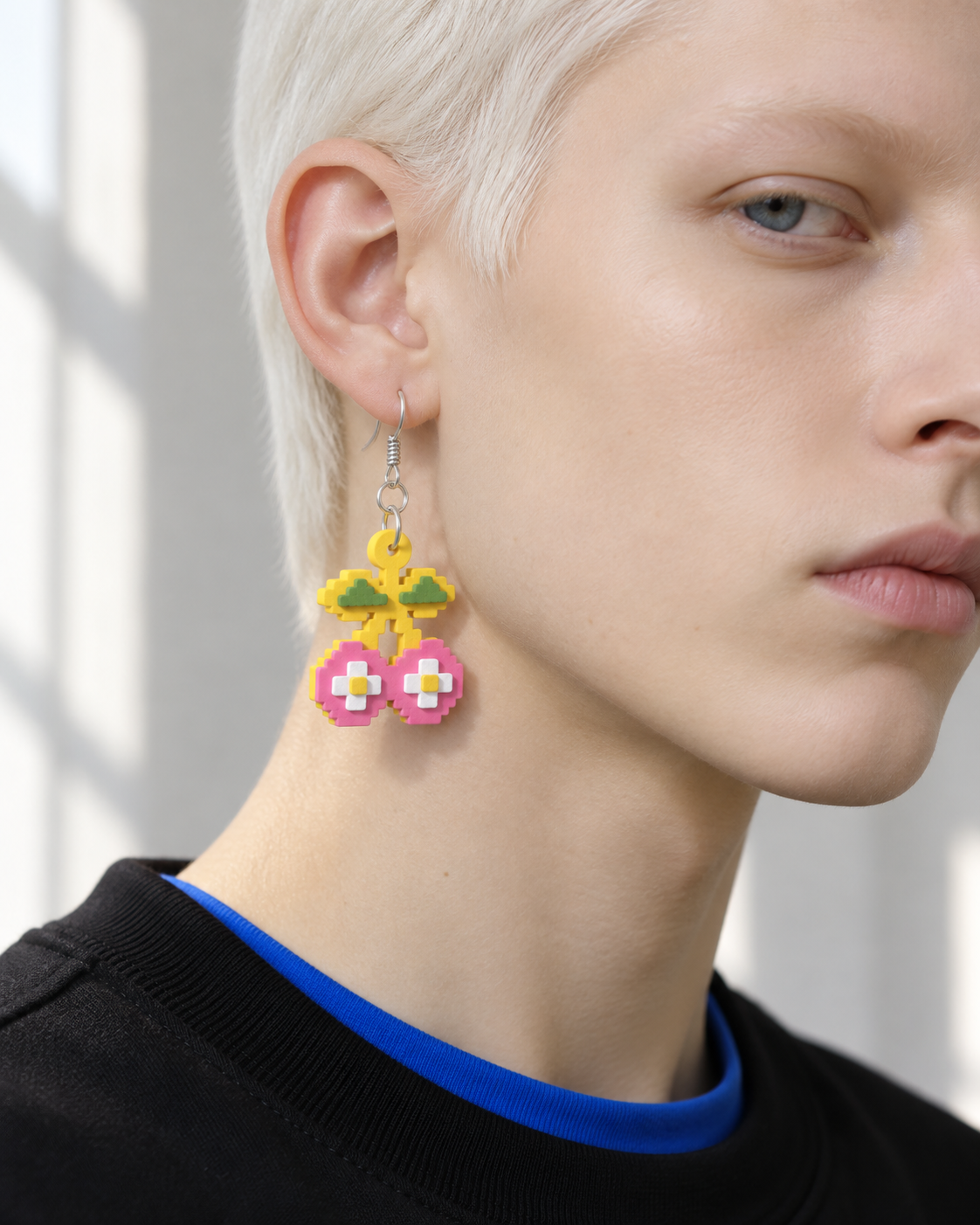 A yellow, pink, green and white Pixel Cherry drop earring worn beside a face, above a blue and black top.