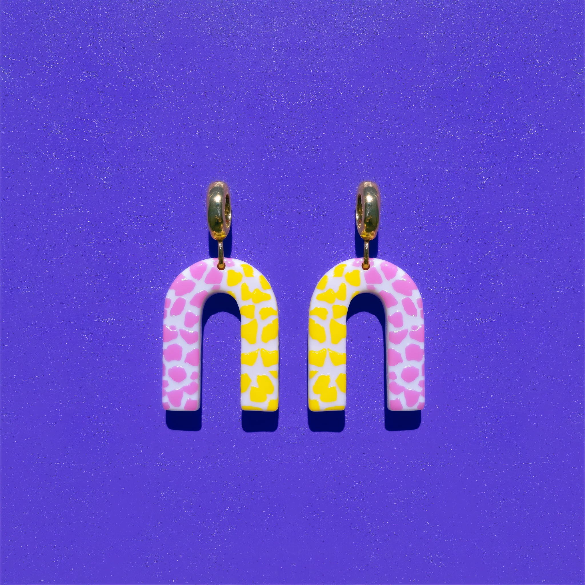 A pair of matching pink and yellow terrazzo arch earrings hang side by side against a violet background.