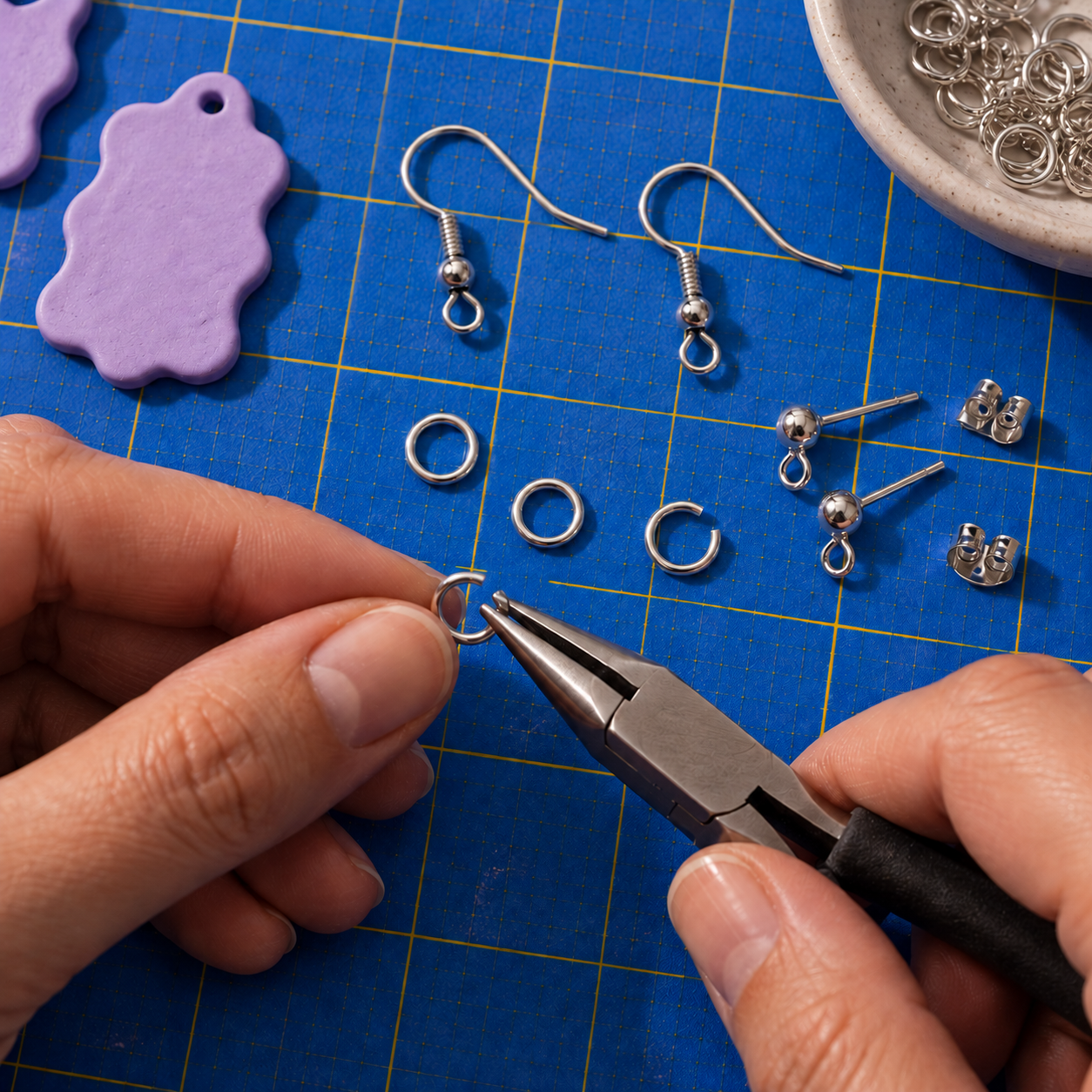Silver ear wires, connecting rings and looped posts on a blue studio mat, beside hands holding a ring with pliers.